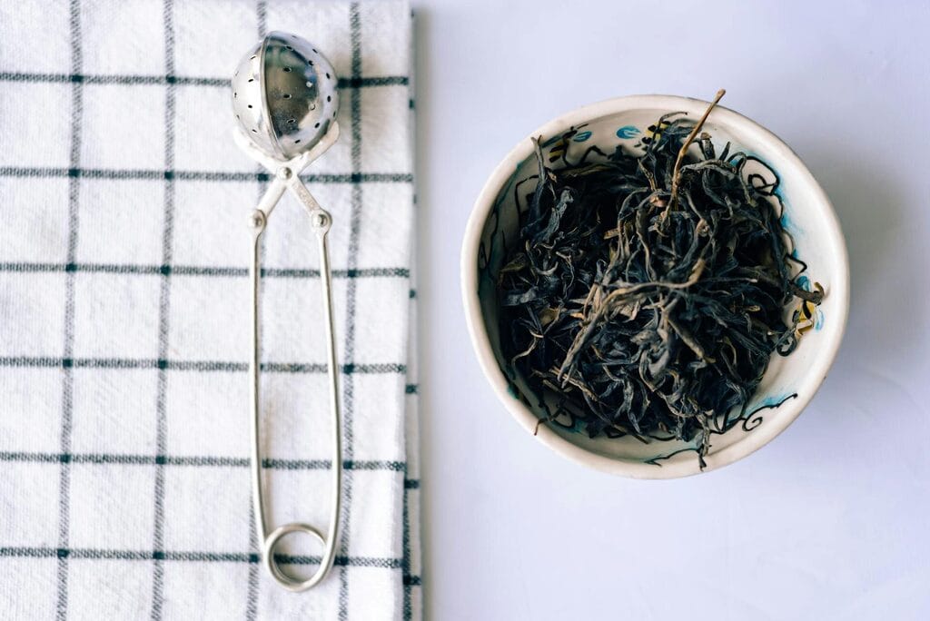 Loose leaf tea in a ceramic bowl beside a vintage tea strainer on a checked linen towel